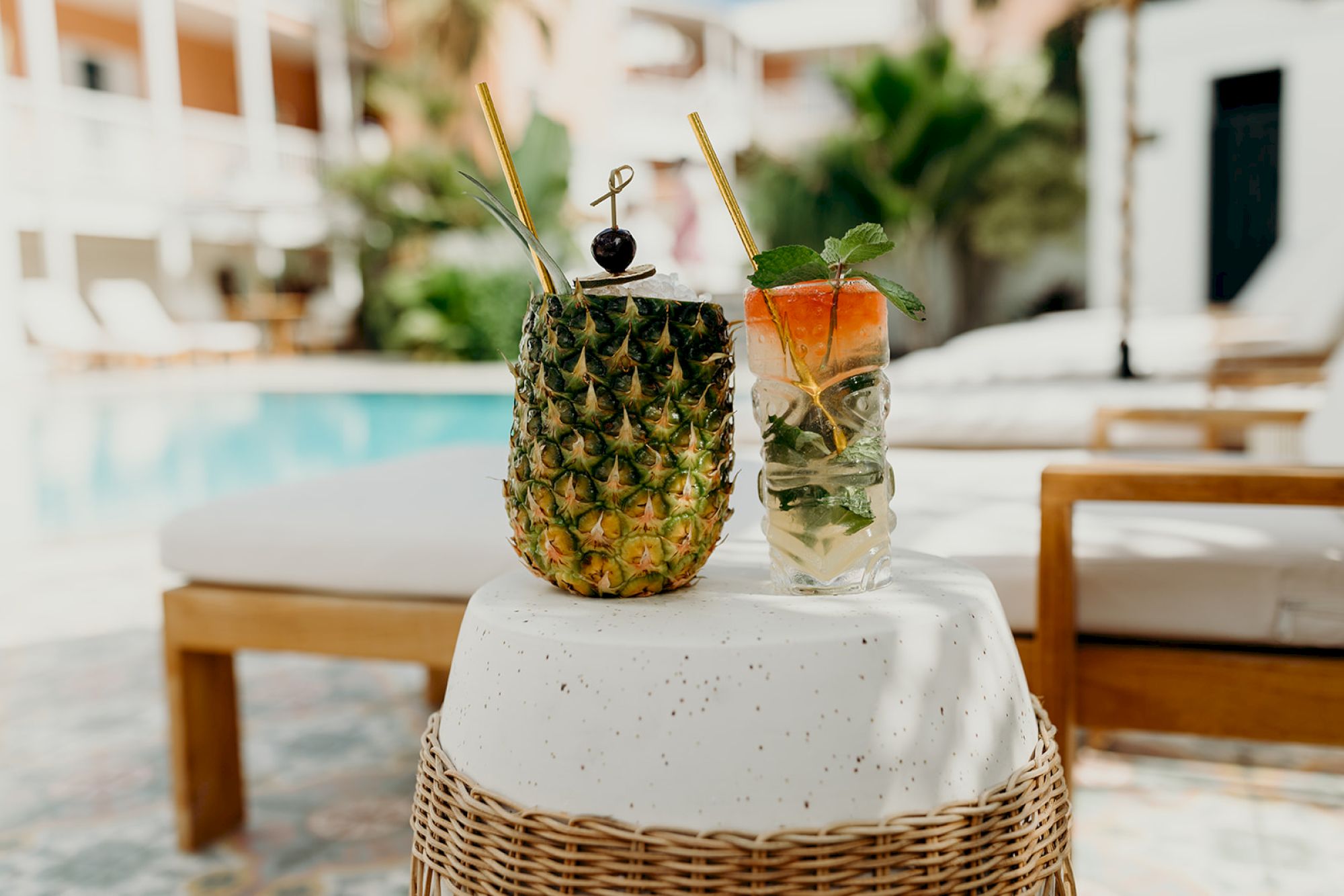 Two drinks, one in a pineapple, the other in a glass with mint, are on a stool by a poolside setting with lounge chairs.