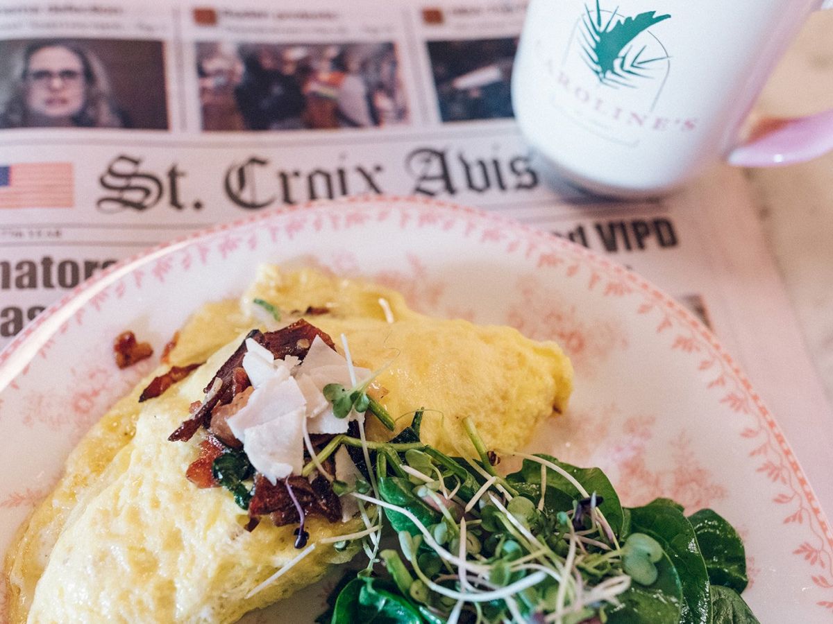 An omelette with greens on a plate, a mug, and a newspaper titled "St. Croix Avis" are on the table.