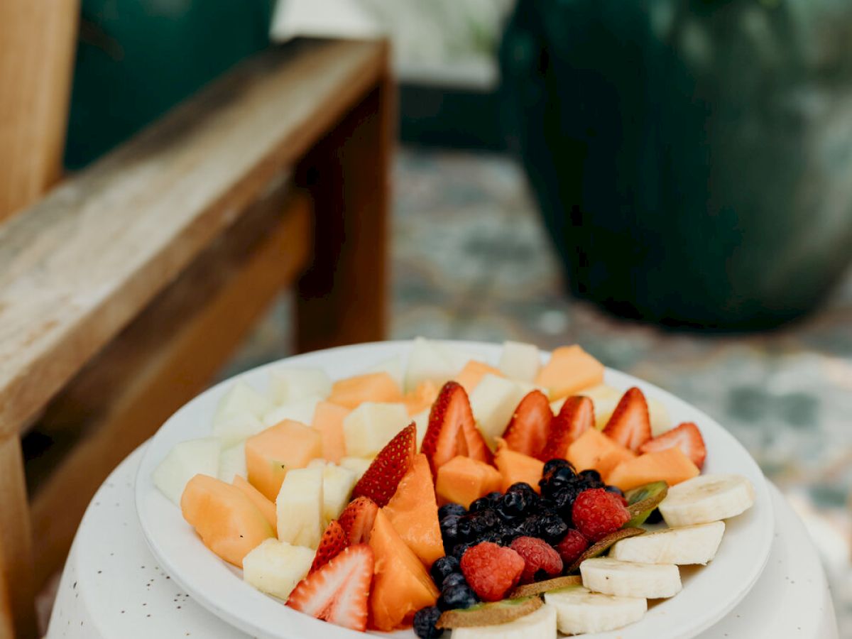 A plate of mixed fruit including strawberries, blueberries, banana slices, and melon sits on a white table outside.