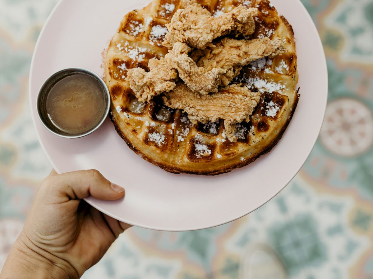 A plate with a waffle topped with something crumbly, and a small cup of syrup on the side, held over a patterned floor.