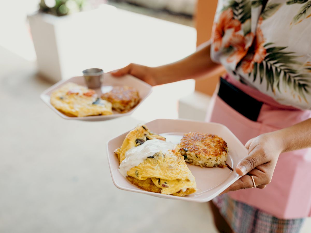 A person is holding two plates with omelets and hash browns, served with a small cup of sauce. They're wearing a floral shirt and pink apron.