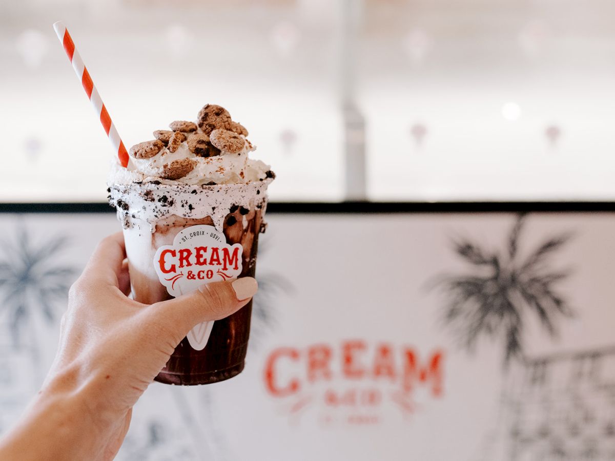 A hand holds a milkshake topped with whipped cream, mini cookies, and a striped straw, in front of a "CREAM & CO" sign with palm tree decor.