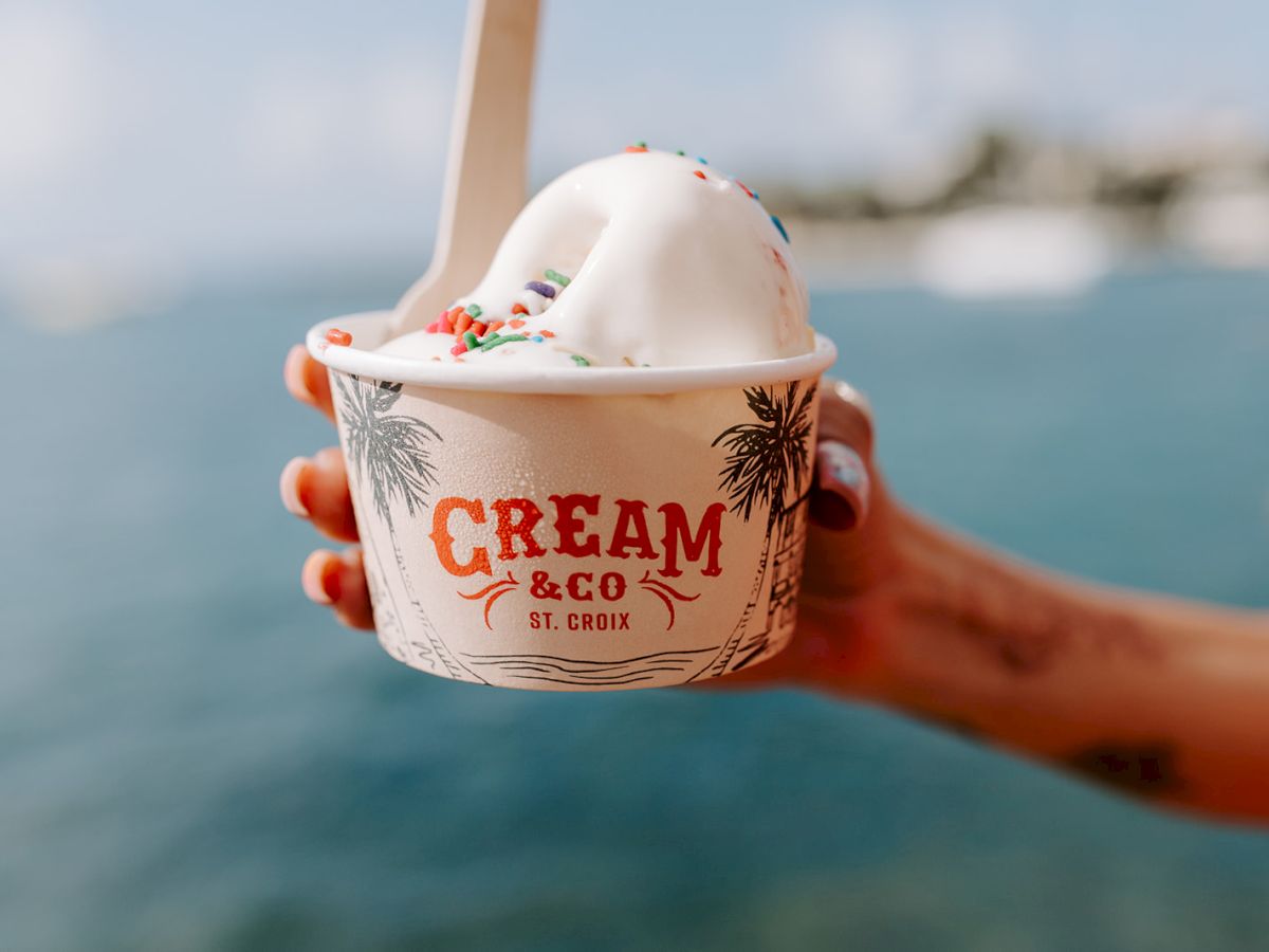 A hand holds a cup of ice cream with sprinkles, labeled "Cream & Co St. Croix," against a blurred waterfront background.