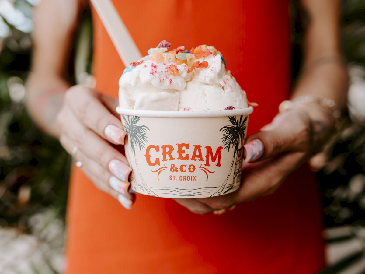 A person holds a cup of ice cream labeled "Cream & Co," topped with colorful toppings, against a blurred background of green foliage.