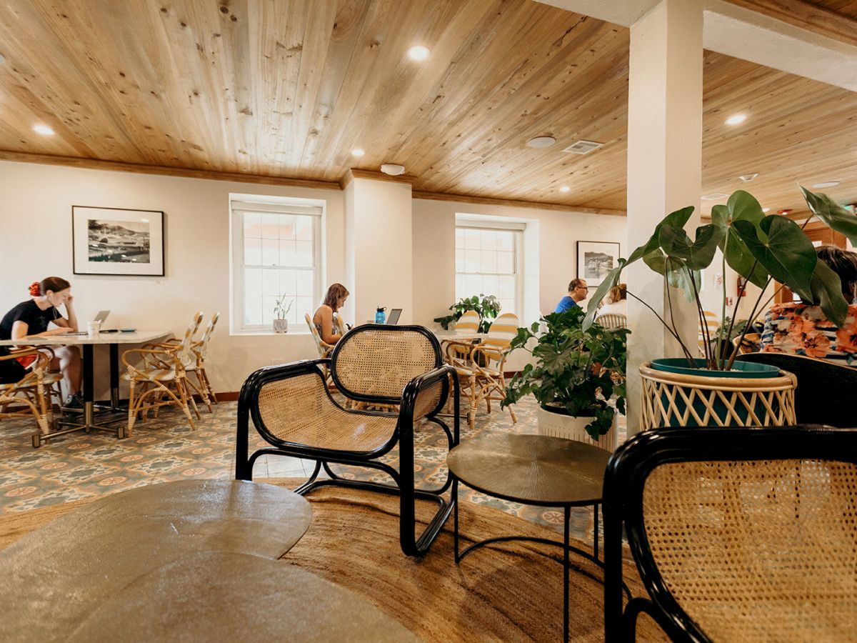 A cozy cafe with people sitting at tables, wooden ceiling, plants, and wicker chairs. Natural light streams through the windows.