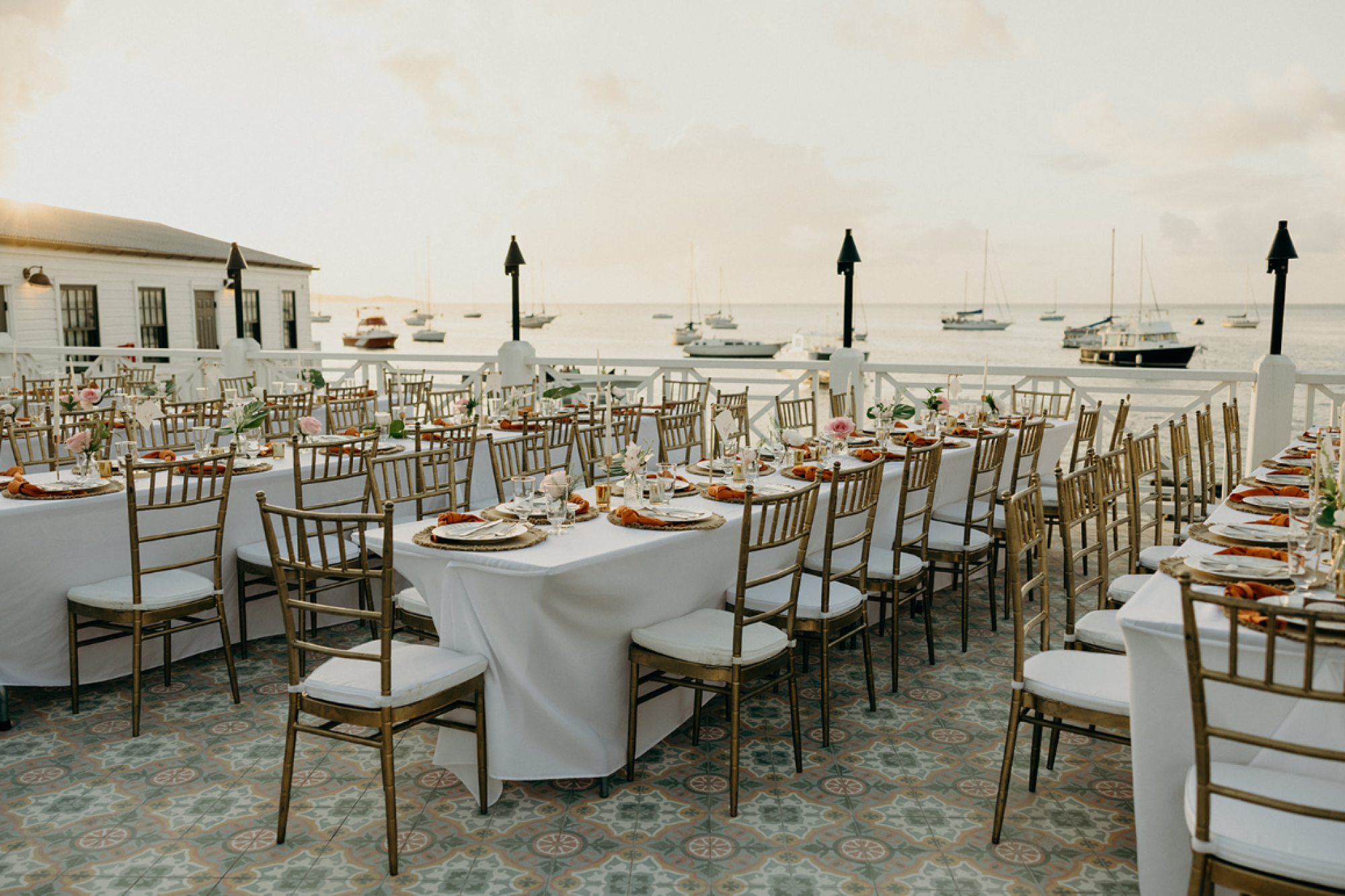 An elegant outdoor dining setup overlooks a harbor with several boats, featuring neatly arranged tables and chairs under a soft sunset glow.