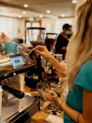 A person is operating an espresso machine in a café, preparing coffee. The environment appears busy and cozy with other people in the background.