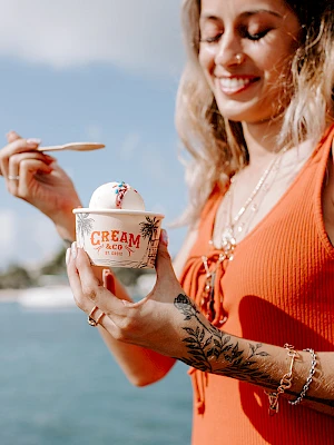 A person in an orange top holds a cup of ice cream by the water, smiling and using a wooden spoon to enjoy it.