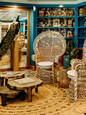 A cozy room with wicker chairs, a peacock, patterned tiles, and bookshelves with a mix of decor and books in a vibrant setting.