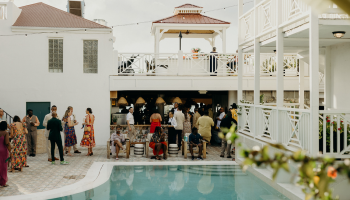 People are gathered around a poolside setting at a social event, with a two-story building and decorative plants in the background.