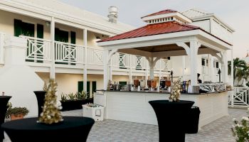 An outdoor bar area with tall cocktail tables, a red-roofed gazebo, and a white building in the background.
