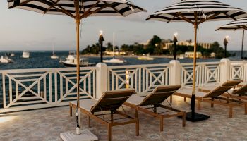 Lounge chairs and striped umbrellas on a patio overlooking a calm sea with boats, near a tree-lined shore under a blue sky.