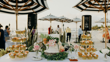 A decorated cake and cupcakes on a table at an outdoor event with umbrellas and people in the background.