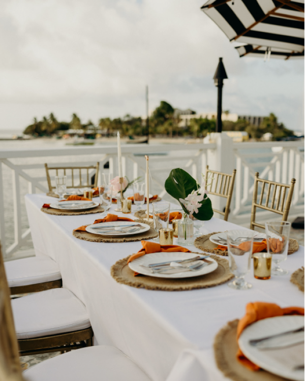 An elegantly set outdoor dining table with white linens, orange napkins, and floral accents, overlooking a calm waterfront view.