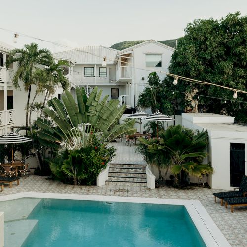 The image shows a serene pool area in a courtyard with surrounding greenery, lounge chairs, and a string of lights.