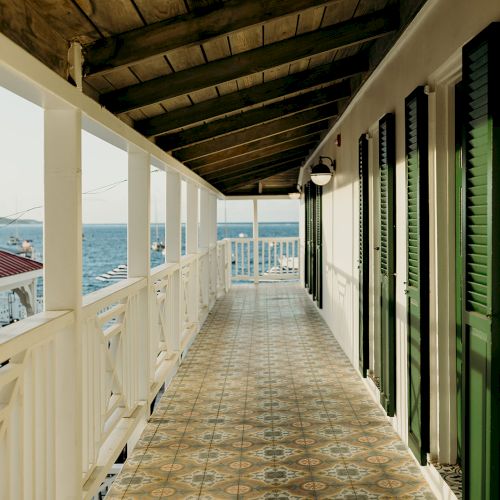 A sunlit veranda with patterned tiles, green shutters, and a wooden roof overlooks a body of water, creating a serene seaside view.