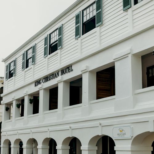 This image shows the exterior of a white building named King Christian Hotel, featuring arched windows and multiple signs at the entrance.