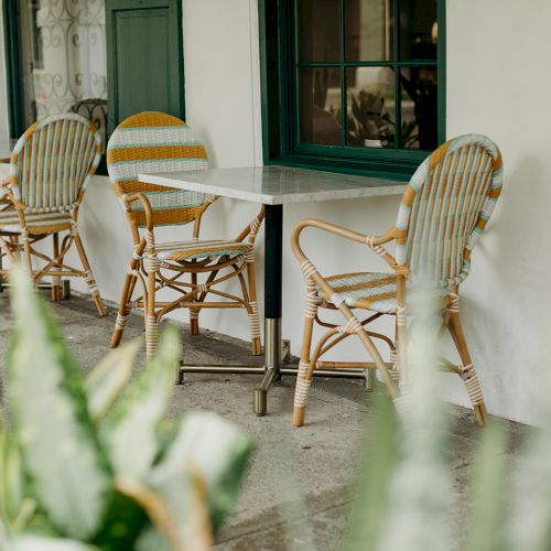 An outdoor seating area with wicker chairs and tables, surrounded by greenery, against a building with green window frames.
