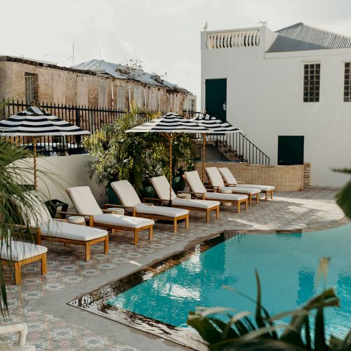 A serene poolside area with lounge chairs, striped umbrellas, and tropical plants. A building is in the background, adding to the tranquil ambiance.