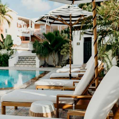 A poolside setting with lounge chairs, umbrellas, and lush greenery surrounded by a building under a sunny sky.