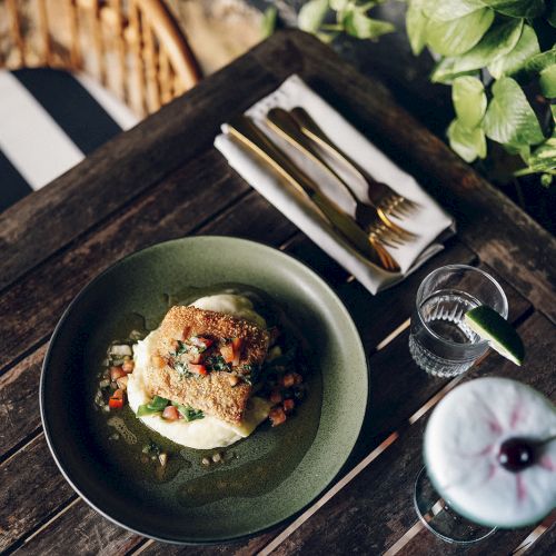A plate with a dish on a wooden table, next to a drink, a glass of water, and cutlery wrapped in a napkin, surrounded by greenery.