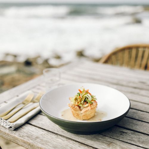 A dish with a garnish on an outdoor table by the sea, with cutlery and glasses, creating a serene dining atmosphere by the water.