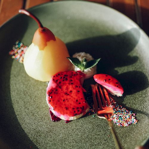 A dessert plate with poached pear, dragon fruit slices, decorative berries, and garnishes on a dark green plate.
