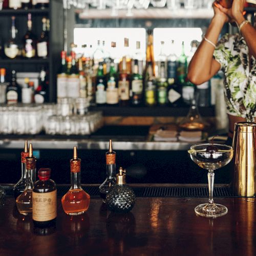 A bartender wearing a mask shakes a cocktail shaker behind a bar, with bottles and cocktail mixers on the counter.