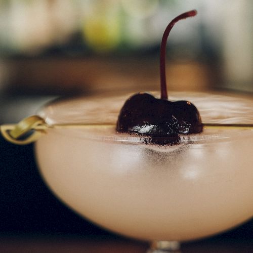 A cocktail with a dark cherry garnish sits in a glass, with a blurred background.