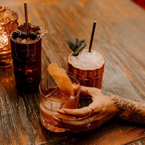 An ornate table with a hand holding a cocktail, alongside two tiki-style drinks and a decorative lantern.