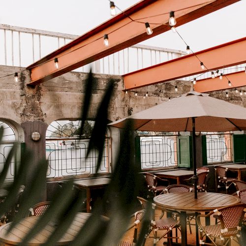 The image shows an outdoor cafe with tables, chairs, umbrellas, string lights, and decorative plants, set in a rustic-looking environment.