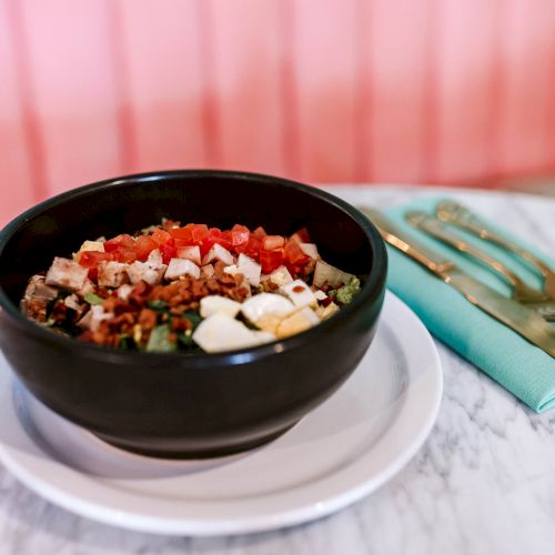 A bowl with a colorful salad, including diced vegetables and what looks like cheese, sits on a table beside wrapped cutlery.