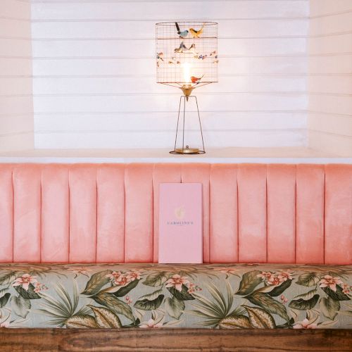 A pink booth with floral patterns, a birdcage lamp, and tropical wallpaper; a menu or book is placed on the seat.