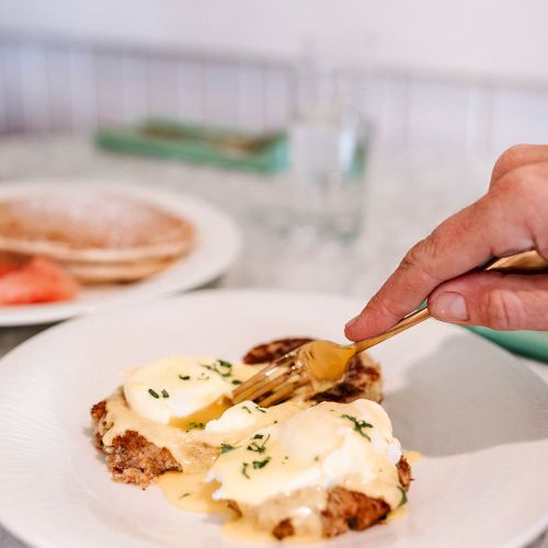 A hand using a fork to cut into eggs Benedict on a white plate, with another plate and a glass in the background.