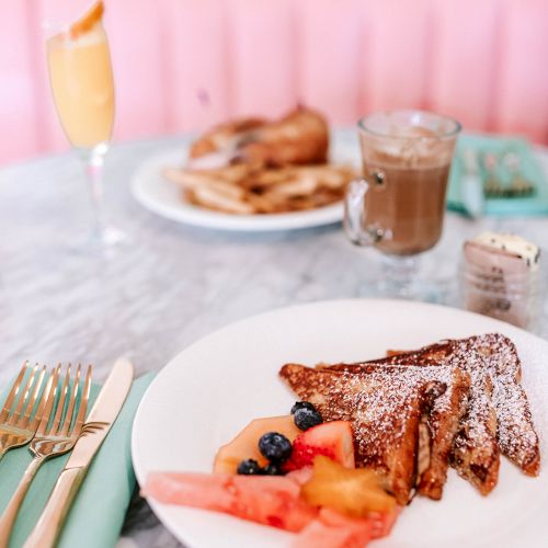 A plate of French toast with fruit, a drink, and coffee on a marble table with a pink background. Another plate is visible in the distance.