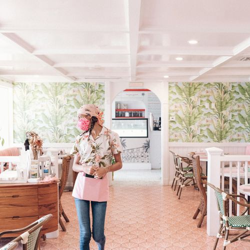 A person walks through a stylish restaurant with patterned walls and tiled floors, carrying items and wearing a mask.