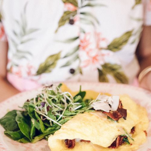 A person holds a plate with an omelette, greens, and sprouts, wearing a floral shirt.