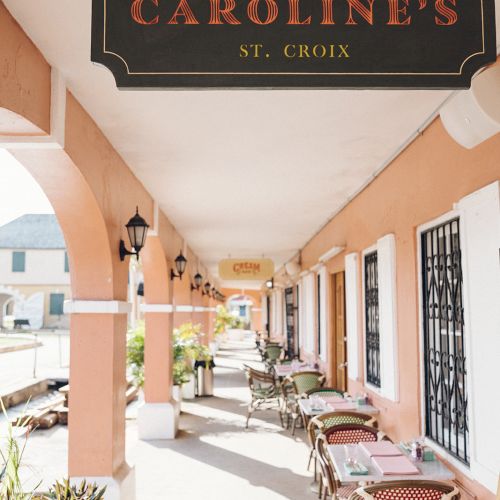 A covered walkway with tables and chairs outside "Caroline's" breakfast and brunch spot in St. Croix, with a sign above.