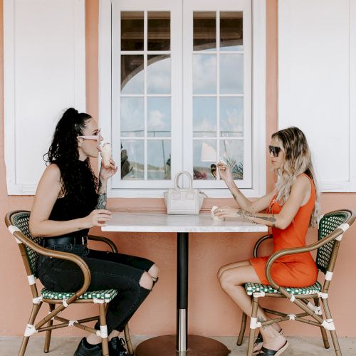 Two people sit at an outdoor table against a peach wall with a window, enjoying drinks and conversation.
