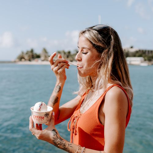 A person is enjoying ice cream by the water, wearing a sleeveless top, with an island visible in the background under a blue sky.