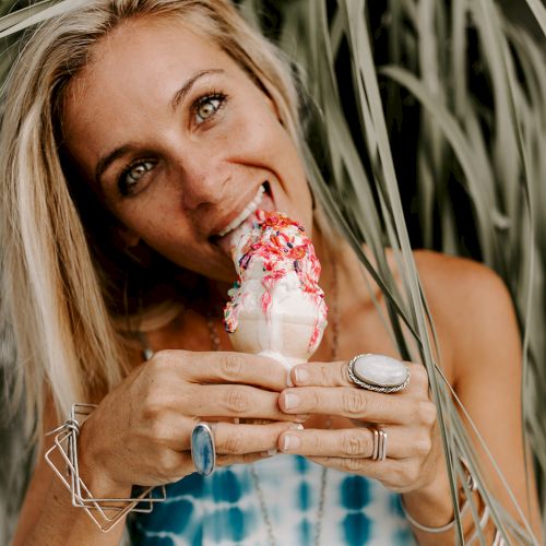 A woman with blonde hair enjoys a soft serve ice cream cone with red syrup, surrounded by green plants, wearing rings and bracelets.