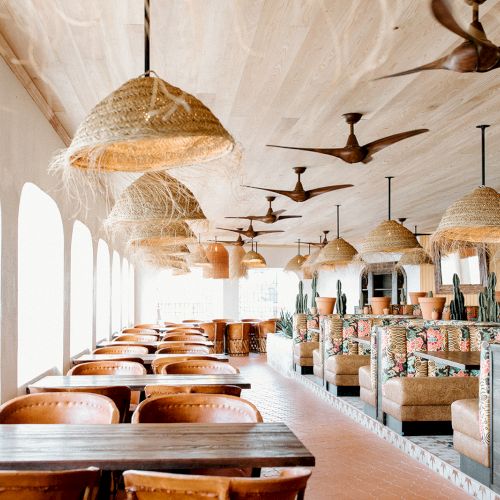The image shows a bright, spacious restaurant with hanging lights, wooden tables, cushioned seating, and ceiling fans.