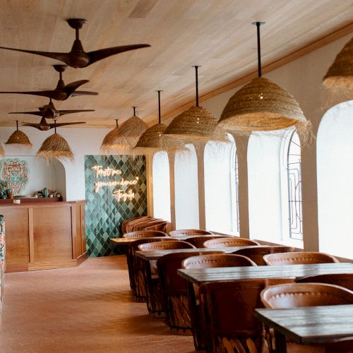 The image shows a stylish, cozy restaurant interior with wooden tables, wicker lamps, decorative tiles, and large arched windows.