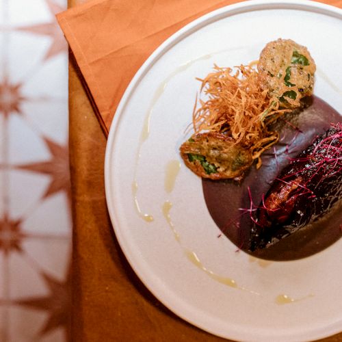 A gourmet dish featuring a stuffed pepper on a dark sauce, garnished with crispy strands and herbs on a white plate.
