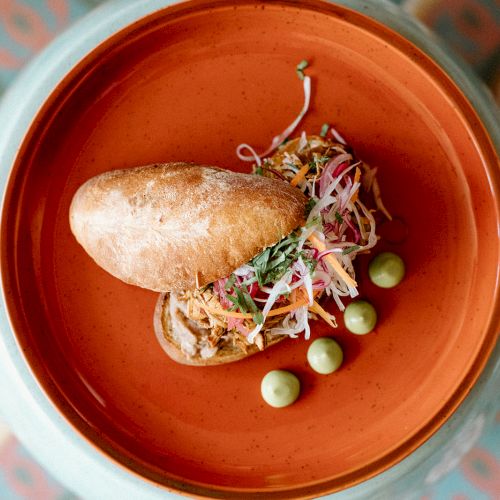 A gourmet dish with a small baguette, colorful salad, sauce, and green pea garnishes on a patterned table.