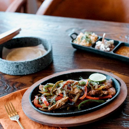 A sizzling platter with meat and vegetables, tortillas in a stone container, plus condiments on a wooden table setting.