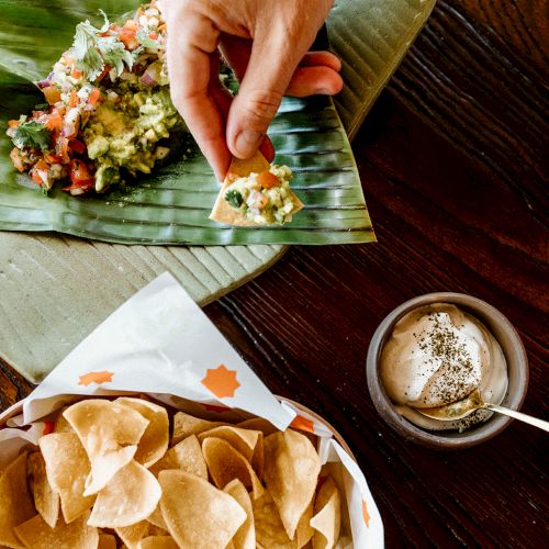 A hand holds a chip with guacamole on a leaf, next to a bowl of chips and a small dish of creamy dip.