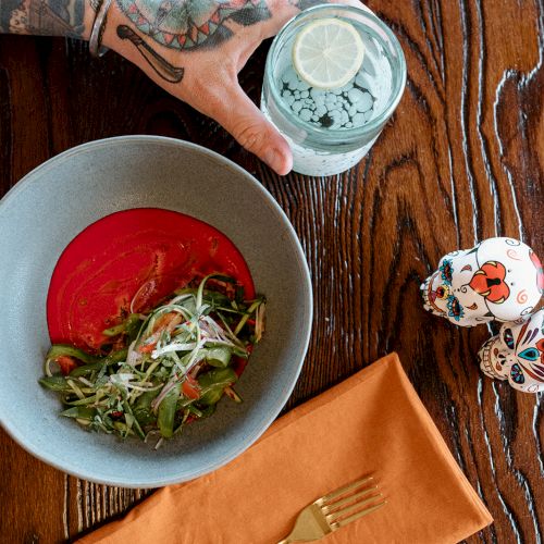 A person with tattoos holds a drink next to a salad in a bowl and a napkin on a wooden table, featuring colorful skull decor.