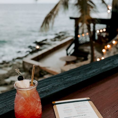 A tropical drink and a menu sit on a table overlooking a beach view with palm trees and ocean in the background.