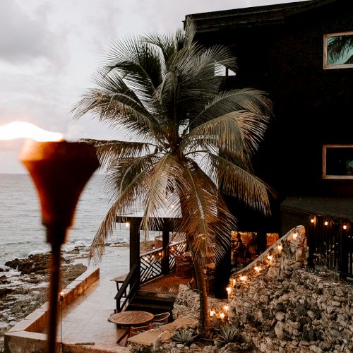 A coastal scene with a wooden house, palm tree, torch, and string lights by the ocean on a rocky shore at sunset.